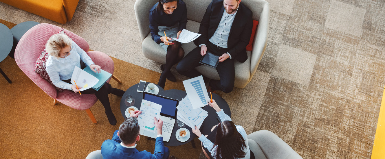 A group of young business people gathered around a coffee table with laptops and paperwork.