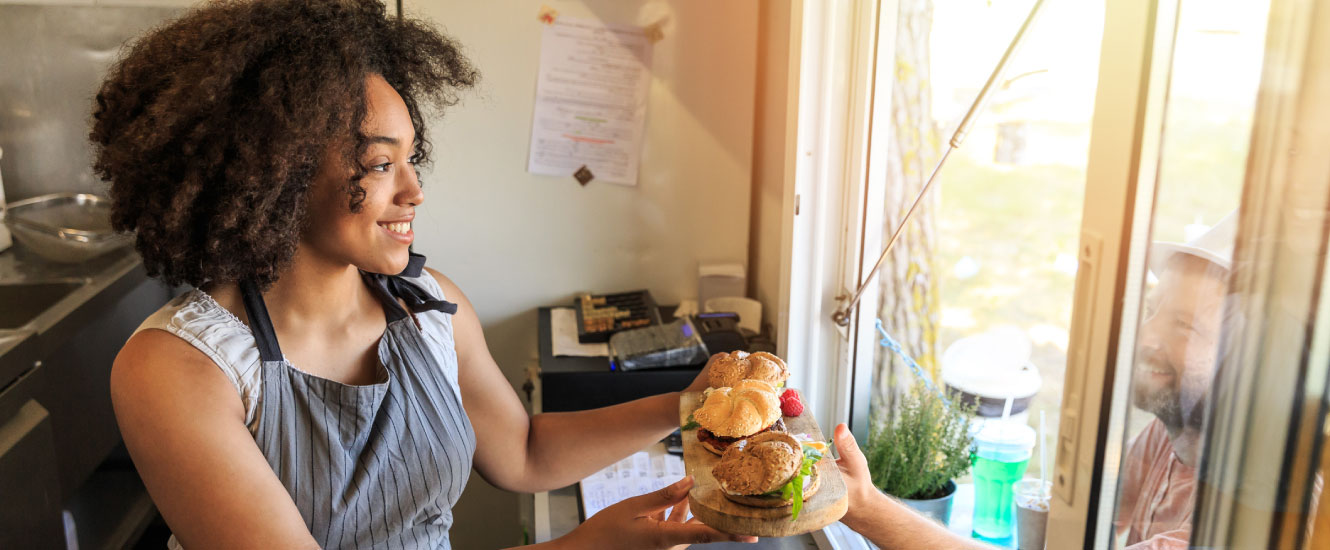 A young woman in an apron holding out a sandwich to a waiting customer.