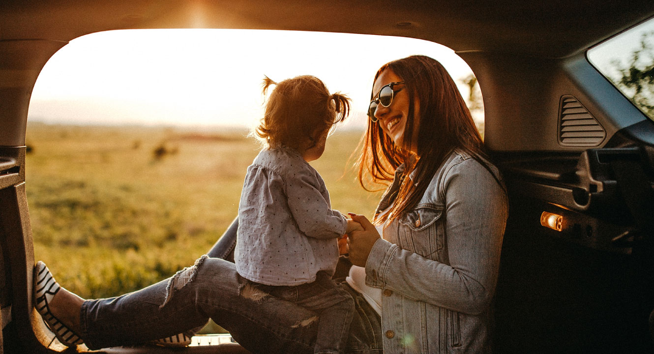 A young mom holding her toddler in the back of a car overlooking a sunset.