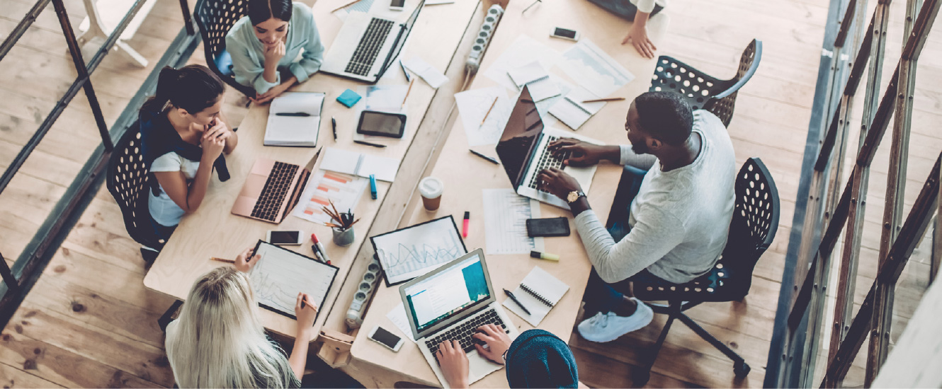 A group of young business people gathered around a large office desk with laptops and paperwork.