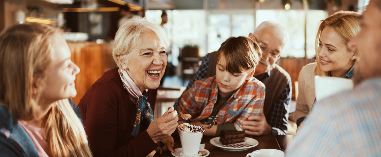 A large happy family gathered around a small tablet inside a coffee shop.