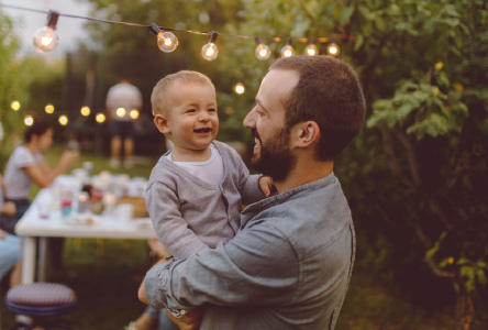 A man holding a small happy child outside in a yard with string lights.