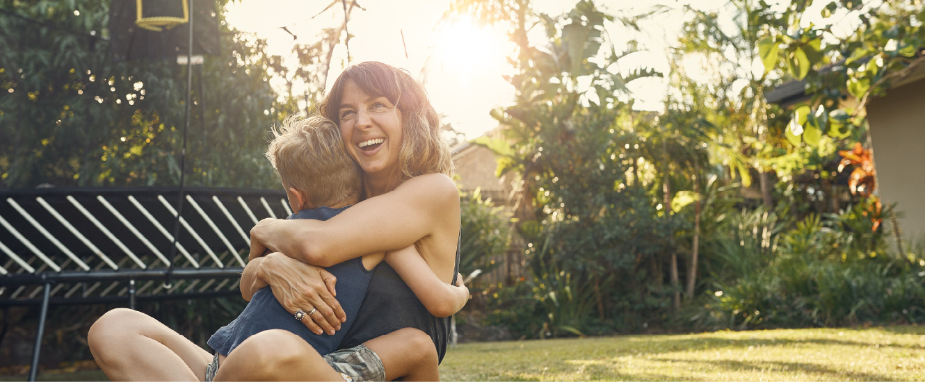 A smiling mom and her young kid hugging outside in a large yard.